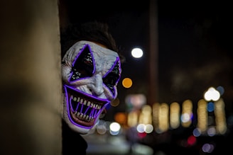 A figure wearing a striking, eerie mask with exaggerated features peeks from behind a wall in a dimly lit setting. The mask is highlighted with purple glowing lines accentuating the eyes and mouth, and sharp teeth are visible. In the background, blurred lights suggest a night-time urban scene.