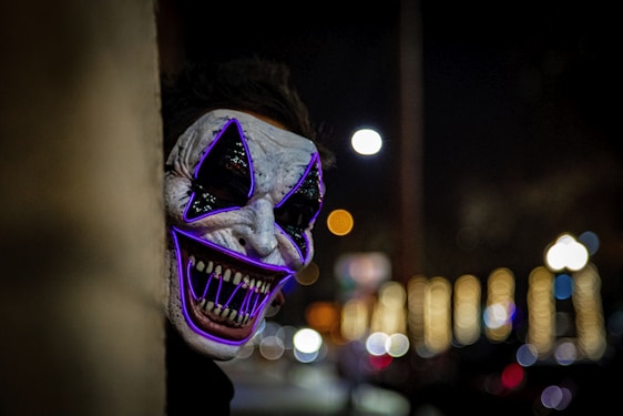 A figure wearing a striking, eerie mask with exaggerated features peeks from behind a wall in a dimly lit setting. The mask is highlighted with purple glowing lines accentuating the eyes and mouth, and sharp teeth are visible. In the background, blurred lights suggest a night-time urban scene.
