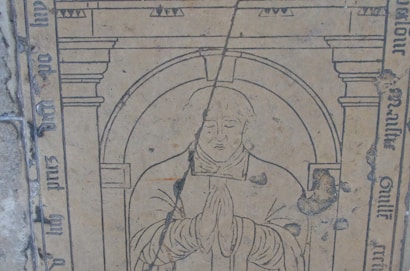 A medieval-style engraving on stone depicting a religious figure in a robe with hands clasped in prayer. The figure is surrounded by decorative arches and Latin script adorns the borders. There is visible wear and damage on the stone, indicating age.