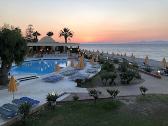 A serene beachside resort at sunset with a large swimming pool surrounded by numerous lounge chairs and yellow umbrellas. Palm trees and a building with an open terrace add to the relaxed atmosphere. The ocean is visible in the background, with calm waves and a horizon tinged with the warm colors of sunset.
