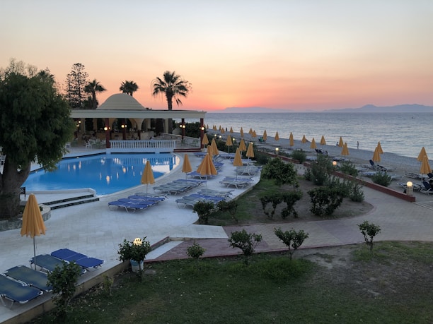 A serene beachside resort at sunset with a large swimming pool surrounded by numerous lounge chairs and yellow umbrellas. Palm trees and a building with an open terrace add to the relaxed atmosphere. The ocean is visible in the background, with calm waves and a horizon tinged with the warm colors of sunset.
