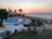 A serene beachside resort at sunset with a large swimming pool surrounded by numerous lounge chairs and yellow umbrellas. Palm trees and a building with an open terrace add to the relaxed atmosphere. The ocean is visible in the background, with calm waves and a horizon tinged with the warm colors of sunset.