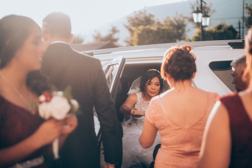 Bride and groom stepping out of a vintage wedding car surrounded by joyful family and friends.