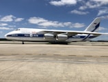 A large cargo aircraft with Volga-Dnepr branding sits on an airport runway. The plane is white with blue accents and features four powerful engines. The sky is clear with scattered clouds, and the ground is a smooth, expansive concrete surface of the airport.