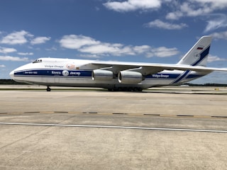 A large cargo aircraft with Volga-Dnepr branding sits on an airport runway. The plane is white with blue accents and features four powerful engines. The sky is clear with scattered clouds, and the ground is a smooth, expansive concrete surface of the airport.