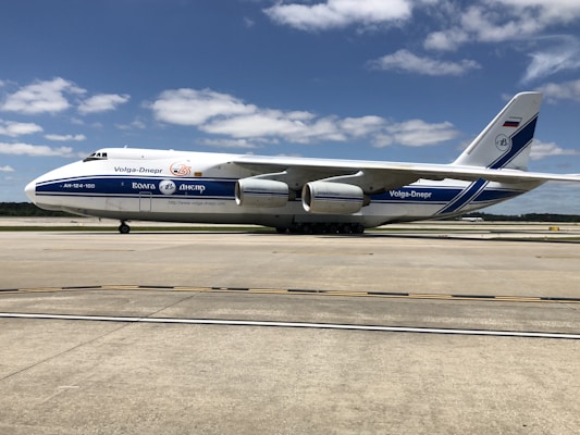 A large cargo aircraft with Volga-Dnepr branding sits on an airport runway. The plane is white with blue accents and features four powerful engines. The sky is clear with scattered clouds, and the ground is a smooth, expansive concrete surface of the airport.