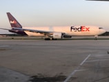 A FedEx cargo plane is stationed on an airport runway, with its logo displayed prominently on the side. The aircraft is large, with a white body and purple accents on the tail fin and engines. The runway appears smooth and expansive, with faint runway markings visible. The background shows a clear sky with a hint of orange, possibly indicating either sunrise or sunset, and some greenery at the horizon.