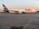 A FedEx cargo plane is stationed on an airport runway, with its logo displayed prominently on the side. The aircraft is large, with a white body and purple accents on the tail fin and engines. The runway appears smooth and expansive, with faint runway markings visible. The background shows a clear sky with a hint of orange, possibly indicating either sunrise or sunset, and some greenery at the horizon.