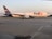 A FedEx cargo plane is stationed on an airport runway, with its logo displayed prominently on the side. The aircraft is large, with a white body and purple accents on the tail fin and engines. The runway appears smooth and expansive, with faint runway markings visible. The background shows a clear sky with a hint of orange, possibly indicating either sunrise or sunset, and some greenery at the horizon.