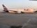 A FedEx cargo plane is stationed on an airport runway, with its logo displayed prominently on the side. The aircraft is large, with a white body and purple accents on the tail fin and engines. The runway appears smooth and expansive, with faint runway markings visible. The background shows a clear sky with a hint of orange, possibly indicating either sunrise or sunset, and some greenery at the horizon.