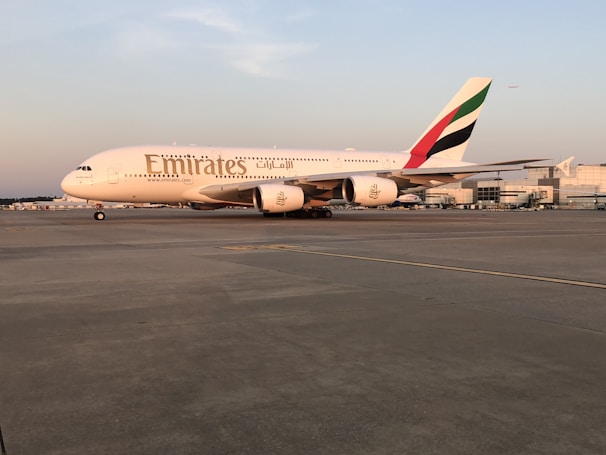 A large Emirates passenger airplane is stationary on an airport runway. The aircraft is primarily white with the Emirates logo displayed prominently on its side and the colors of the United Arab Emirates flag on the tail fin. The sun appears to be setting or rising, casting a warm hue over the plane and the runway. In the distance, airport buildings and another small aircraft are visible.