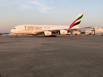 A large Emirates passenger airplane is stationary on an airport runway. The aircraft is primarily white with the Emirates logo displayed prominently on its side and the colors of the United Arab Emirates flag on the tail fin. The sun appears to be setting or rising, casting a warm hue over the plane and the runway. In the distance, airport buildings and another small aircraft are visible.