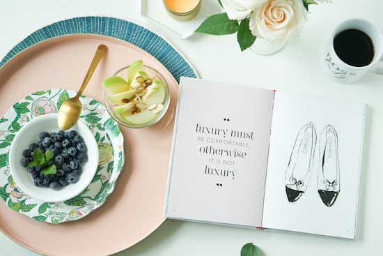 On a white surface, there is a flat lay arrangement featuring a pink tray. Inside the tray, there's a small floral plate with a bowl of blueberries and a mint leaf. Next to the bowl is a glass cup filled with sliced green apples and almonds. An open book displays a quote on one page and a sketch of ballet flats on the other. Nearby, a white mug contains black coffee and a white candle burns beside a vase of white roses.