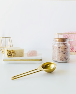 A minimalistic arrangement featuring a bar of soap with floral embellishments, a pink crystal, and a jar of pink bath salts on a white tray. A golden measuring spoon is placed in the foreground, with geometric candle holders and a pink towel in the background.
