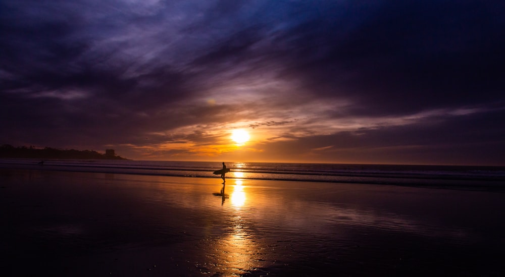 silhouette photography of person standing on shore while holding surfboard