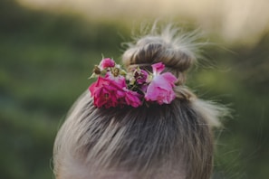 A hairstyle featuring a bun adorned with vibrant pink flowers, set against a blurred green background. The flowers add a touch of natural beauty and contrast with the hair color.
