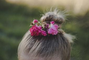 A hairstyle featuring a bun adorned with vibrant pink flowers, set against a blurred green background. The flowers add a touch of natural beauty and contrast with the hair color.