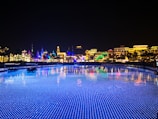 A vibrant and colorful night scene of a theme park or entertainment area with illuminated buildings showcasing various cultural designs. The foreground features a body of water with blue LED lights creating a sparkling effect, reflecting the bright lights of the architecture.