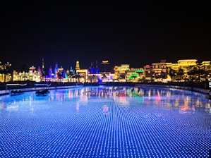 A vibrant and colorful night scene of a theme park or entertainment area with illuminated buildings showcasing various cultural designs. The foreground features a body of water with blue LED lights creating a sparkling effect, reflecting the bright lights of the architecture.