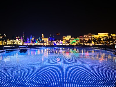 A vibrant and colorful night scene of a theme park or entertainment area with illuminated buildings showcasing various cultural designs. The foreground features a body of water with blue LED lights creating a sparkling effect, reflecting the bright lights of the architecture.