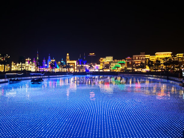 A vibrant and colorful night scene of a theme park or entertainment area with illuminated buildings showcasing various cultural designs. The foreground features a body of water with blue LED lights creating a sparkling effect, reflecting the bright lights of the architecture.