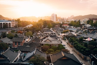 A picturesque view of a traditional Korean village with numerous tiled-roof houses surrounded by lush greenery. In the background, modern buildings and a sunset sky create a blend of tradition and modernity.