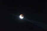 A celestial body is partially illuminated against a dark night sky, creating a striking contrast between the bright white section and the shadowed area. Two beams of light radiate from the bright part, enhancing the dramatic effect.