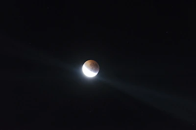 A celestial body is partially illuminated against a dark night sky, creating a striking contrast between the bright white section and the shadowed area. Two beams of light radiate from the bright part, enhancing the dramatic effect.