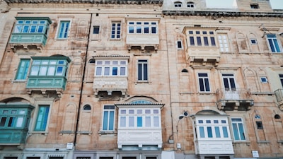 A facade of an old, multi-story building with large rectangular wooden balconies painted in shades of green and cream. Some windows are arched while others are rectangular, adding architectural variety. The outer walls are built with light brown stone, showing signs of aging with worn patches and cracks.