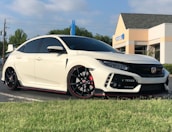 A sleek white Honda car with black and red accents is parked on a street next to a grassy patch. The vehicle has a sporty design with custom rims and tinted windows. In the background, there is a Chase bank building with clear blue skies above.