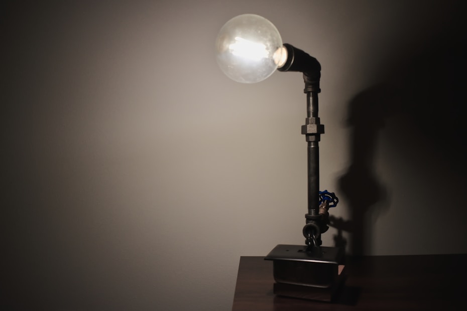 A table lamp made from industrial materials with a visible bulb on top. The lamp appears to be constructed from metal pipes and fittings, casting a shadow on the wall behind it. It is placed on a wooden surface.