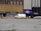 A street scene features a white and yellow ambulance parked in front of a pizza restaurant with a purple awning. The restaurant is situated between two buildings, one with brown tiled facade and the other with an eye care sign. A beige sedan is also parked nearby. The street is lined with concrete barriers and traffic cones, and the pavement shows signs of wear with visible cracks.