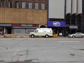 A street scene features a white and yellow ambulance parked in front of a pizza restaurant with a purple awning. The restaurant is situated between two buildings, one with brown tiled facade and the other with an eye care sign. A beige sedan is also parked nearby. The street is lined with concrete barriers and traffic cones, and the pavement shows signs of wear with visible cracks.