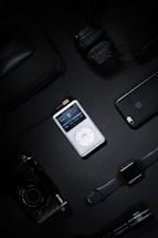 A collection of electronic gadgets including a classic iPod, over-ear headphones, a smartphone, a digital camera, a smart watch, and a computer mouse arranged on a dark surface.