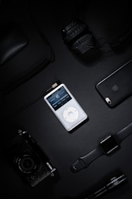 A collection of electronic gadgets including a classic iPod, over-ear headphones, a smartphone, a digital camera, a smart watch, and a computer mouse arranged on a dark surface.