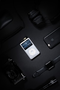 A collection of electronic gadgets including a classic iPod, over-ear headphones, a smartphone, a digital camera, a smart watch, and a computer mouse arranged on a dark surface.
