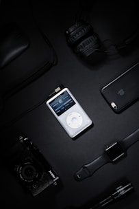 A collection of electronic gadgets including a classic iPod, over-ear headphones, a smartphone, a digital camera, a smart watch, and a computer mouse arranged on a dark surface.