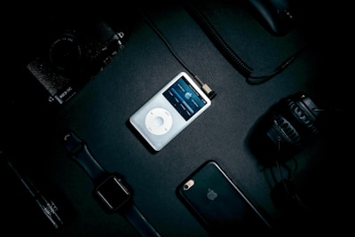 A collection of electronic devices arranged on a dark surface includes an iPod with its screen lit up in the center. Surrounding it are a smartwatch, a smartphone with an Apple logo, a digital camera, headphones, and various cables, creating a tech-centric still life.