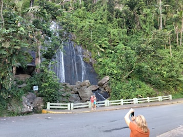 A lush green landscape with tall, dense trees and a small waterfall cascading over dark rocks. A person on the road is taking a photo of another person posing near the waterfall. The scene is vibrant with greenery surrounding the waterfall and rocky area.