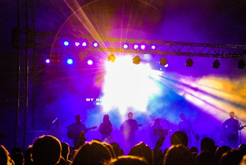 A lively concert scene with a band performing on stage, silhouetted against bright and colorful stage lights. The audience is gathered in front, with hands raised, creating a dynamic and energetic atmosphere. Purple and yellow lights dominate the background, adding vibrancy to the scene.