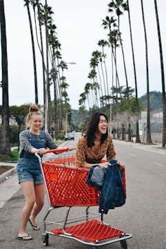 Two women are having fun on a palm-lined street, with one woman playfully pushing a shopping cart containing the other, who is laughing joyfully. Both are dressed casually; the woman in the cart is wearing a patterned top, and the one pushing wears denim overalls. The vibrant red shopping cart stands out against the overcast sky and the tall palm trees lining the road.