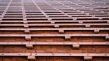 Wooden bleachers expand across the image, showcasing a series of evenly spaced wooden benches set against a stone backdrop. The angular and repeating pattern creates a sense of uniformity and order.