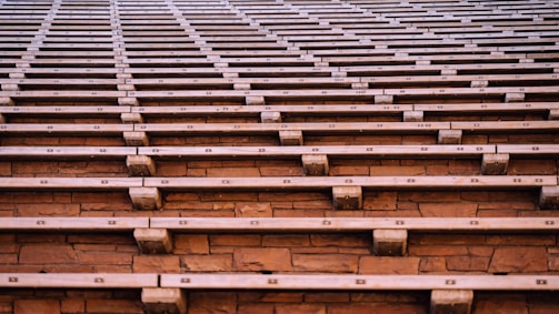 Wooden bleachers expand across the image, showcasing a series of evenly spaced wooden benches set against a stone backdrop. The angular and repeating pattern creates a sense of uniformity and order.