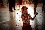 A young girl joyfully playing with flower petals that are gently falling around her. She is wearing colorful traditional clothing and appears to be in a festive or celebratory environment, with people in the background. The light creates a soft, warm glow, enhancing the scene's atmosphere.
