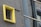 A small, square window with metal bars is surrounded by a bright yellow frame on a gray concrete wall. Adjacent is a reflective glass surface, possibly part of a modern building, which contrasts with the rough texture of the wall.