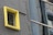 A small, square window with metal bars is surrounded by a bright yellow frame on a gray concrete wall. Adjacent is a reflective glass surface, possibly part of a modern building, which contrasts with the rough texture of the wall.