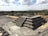 A large ancient stepped pyramid is situated in an open area surrounded by trees and distant hills. The structure is made of stone and features numerous steps leading to its top. A few people are walking and gathering around the base of the pyramid. The sky is partly cloudy.
