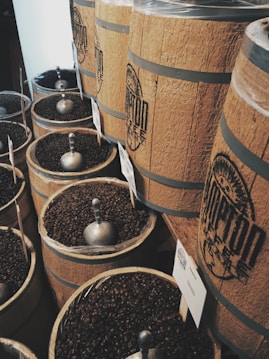 Large wooden barrels filled with dark roasted coffee beans are lined up, each with a metal scoop resting on top. The barrels are branded with a logo, and labels are attached, possibly indicating different types of coffee.