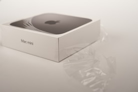A Mac mini box is displayed with its sleek design, partially covered by transparent plastic wrapping. The box is primarily white, featuring the Apple logo and branding on the top.