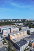 Aerial view of an industrial or office complex with multiple brick buildings featuring large windows. The area is surrounded by greenery and residential neighborhoods, with hills visible in the distance under a clear blue sky.
